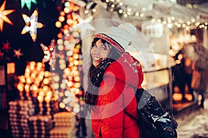 Beautiful young smiling woman enjoy snow winter time on Christmas fair in night city wearing hat and red jacket.