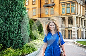 Beautiful young smiling woman walking in city park near famous old architecture building at summer day wearing dress and looking