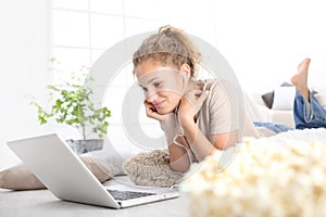 beautiful young smiling woman watch a movie at the computer eating popcorn lying on living room wooden floor in comfortable home