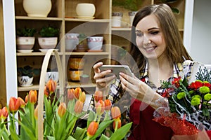 Beautiful young smilling woman florist are taking a picture on her smartphone in flower shop