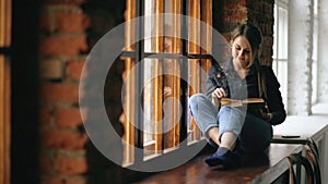 Beautiful young student girl reading book sit on windowsill in university classroom indoors