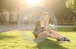Beautiful young student girl sitting on the lawn in the university park reading the book. Cheerful female student sitting on the