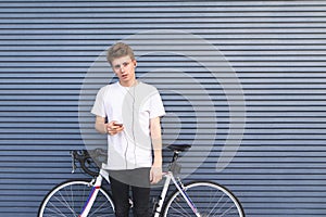 Beautiful young student standing with a smartphone in his hands and headphones near a bicycle on the background