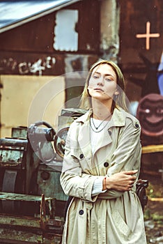 Beautiful young stylish blonde woman wearing long beige coat, white boots, and backpack posing through the city streets. Trendy