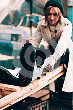 Beautiful young stylish blonde woman wearing long beige coat, white boots, black hat posing through the city streets. Trendy