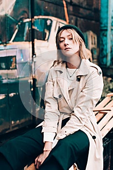 Beautiful young stylish blonde woman wearing long beige coat, white boots, black hat posing through the city streets. Trendy