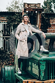 Beautiful young stylish blonde woman wearing long beige coat, white boots and black hat posing on the old green truck. Trendy