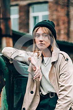 Beautiful young stylish blonde woman wearing long beige coat, white boots and black hat posing on the old green truck. Trendy