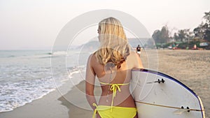 A beautiful young surfer woman is walking along the beach with a surfboard