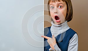 Beautiful young surprised girl looking behind white empty banner. Free copy space