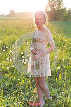 Beautiful young sweet girl in a white dress with hair on the head oblique walks in a field at sunset