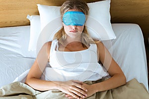 Beautiful young tired woman trying to sleep with sleep mask resting on bed in bedroom at home.