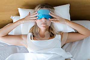 Beautiful young tired woman trying to sleep with sleep mask resting on bed in bedroom at home.