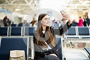 Beautiful young tourist girl in international airport, taking funny selfie with passport and boarding pass near flight information