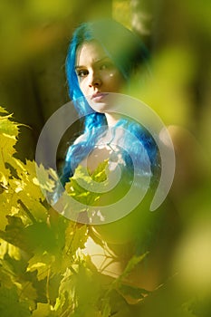 Beautiful young unconventional woman Emo with individual blue hairs, peers out in autumn forest between green yellow leaves.