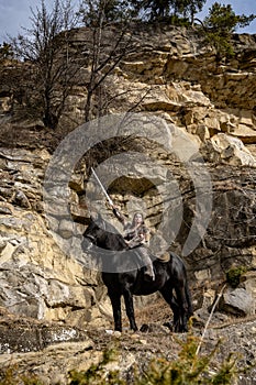 Beautiful young viking warrior woman with sword and big black horse in the mountains scouting the horizont