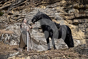 Beautiful young viking warrior woman with sword and big black horse in the mountains scouting the horizont