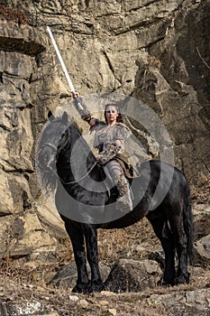 Beautiful young viking warrior woman with sword and big black horse in the mountains scouting the horizont photo