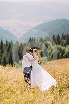 Beautiful young wedding couple walking on sunny field with magnificent forest hills as background. Back view