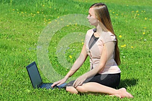 A beautiful young white girl in a pink jacket and black skirt and with long hair sitting on green grass, on the lawn and working