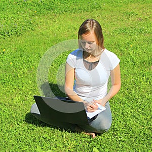 A beautiful young white girl in a white t-shirt and with long hair sitting on green grass, on the lawn and working behind a black