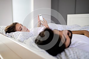 Beautiful young wife texting with lover on smartphone while husband is sleeping