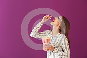 Beautiful young woman with 3D cinema glasses eating popcorn on color background