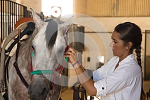 Beautiful young woman adjusting her horse's chinstrap inside a stable. Concept horse riding, animals, saddle, horsewoman