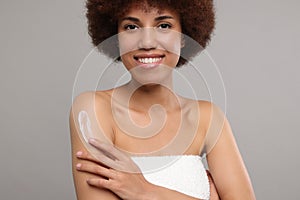 Beautiful young woman applying body cream onto arm on grey background