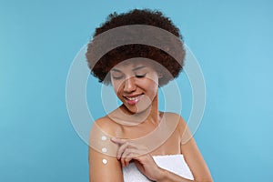 Beautiful young woman applying body cream onto arm on light blue background