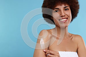 Beautiful young woman applying body cream onto arm on light blue background, space for text