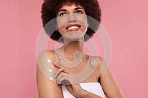 Beautiful young woman applying body cream onto arm on pink background