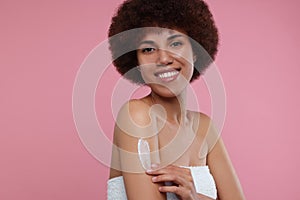 Beautiful young woman applying body cream onto arm on pink background, space for text