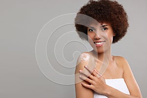 Beautiful young woman applying body cream onto shoulder on grey background, space for text
