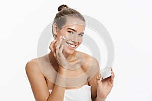 Beautiful young woman applying cosmetic cream treatment on her face isolated on white background