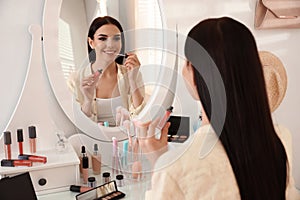 Beautiful young woman applying makeup near mirror indoors