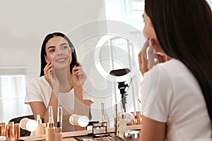 Beautiful young woman applying makeup at table with mirror and ring lamp