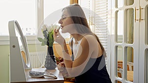 Beautiful young woman applying red lipstick while sitting at makeup table next to big window in bedroom