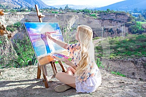 Beautiful young woman artist paints a landscape in nature. Drawing on the easel with colorful paints in the open air.