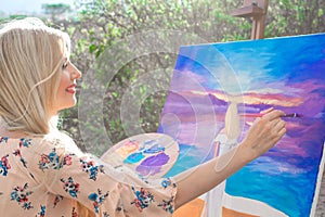 Beautiful young woman artist paints a landscape in nature. Drawing on the easel with colorful paints in the open air.