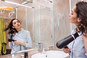 Beautiful young woman in bath towel is using a hair dryer and looking into the mirror