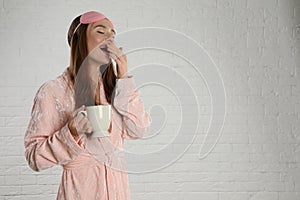 Beautiful young woman in bathrobe and eye sleeping mask yawning near white wall. Space for text