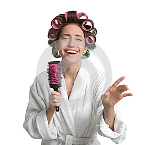 Beautiful young woman in bathrobe with hair curlers singing into hairbrush on white background