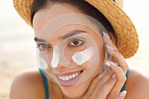 Beautiful young woman at beach applying sunscreen on face and looking at camera. Beauty girl applying suntan lotion at sea