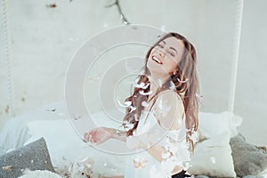 Beautiful young woman in a bed dropping the feathers and smiling. Close-up horizontal portrait.