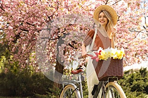 Beautiful young woman with bicycle and flowers in park on pleasant spring day. Space for text