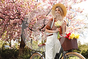 Beautiful young woman with bicycle and flowers in park on pleasant spring day. Space for text