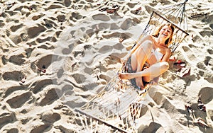 Beautiful young woman in bikini top swinging in hammock on the sandy beach and cheerful laughing in camera top view