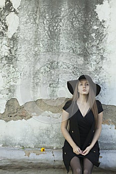 Beautiful young woman in a black dress and a black hat against the old wall
