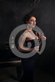 Beautiful young woman in black dress next to a piano with candelabra candles and wine, dark dramatic atmosphere of the castle.